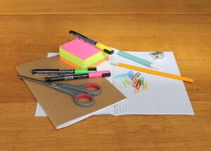 Fournitures scolaires sur table en bois : cahier à carreaux ouvert, post-it fluo, stylos NAGA, surligneur, crayon, taille-crayon, ciseaux gris, trombones.
