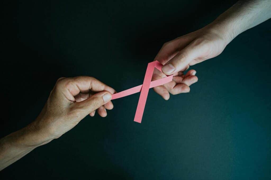 Deux mains se passent un ruban rose de sensibilisation au cancer, sur un fond sombre et uni, symbolisant le soutien et le partage.